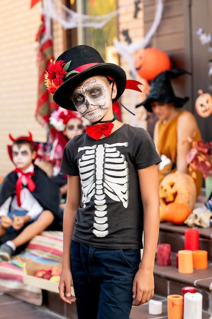 Foto leuke jongen met geschilderd gezicht in t-shirt met skelet en elegante hoed die voor de camera staat tegen zijn vrienden en jonge vrouw