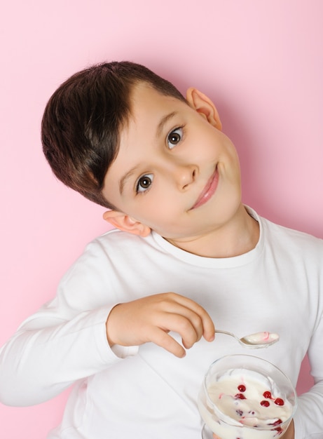 Leuke jongen van 6 jaar oud in een wit t-shirt dat bessen en yoghurt eet uit een glazen kom op een roze ondergrond