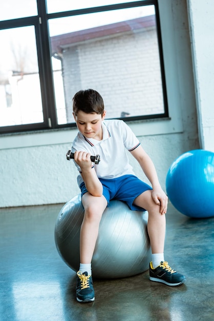 Leuke jongen zittend op een fitnessbal en trainen met een halter in de sportschool