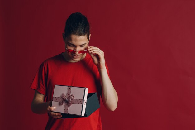 Leuke kerel met geschenkdoos op rode achtergrond. Jonge man met kerst geschenkdoos aanwezig. Leuke verraste jongen op rode achtergrond