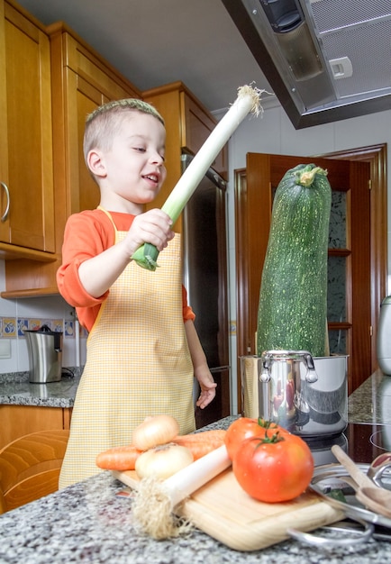 Leuke kindchef-kok met schort die grote courgette en andere groenten in een pot op de keuken kookt