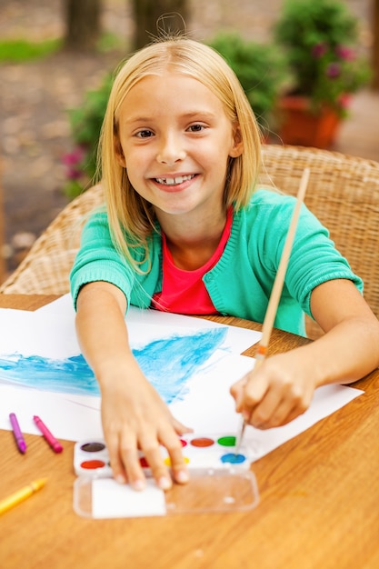 Leuke kleine kunstenaar. Schattig klein meisje dat iets op papier tekent en glimlacht terwijl ze aan tafel en buiten zit