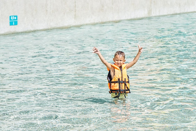 Leuke kleuterjongen in zwemvest staat in zwembad met opgeheven handen in waterpark