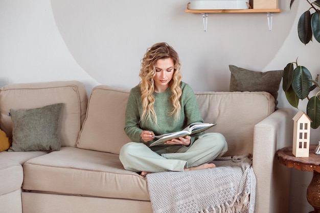 Foto leuke, krullende blonde vrouw leest een boek thuis zittend op de bank student meisje voorbereiding op examens