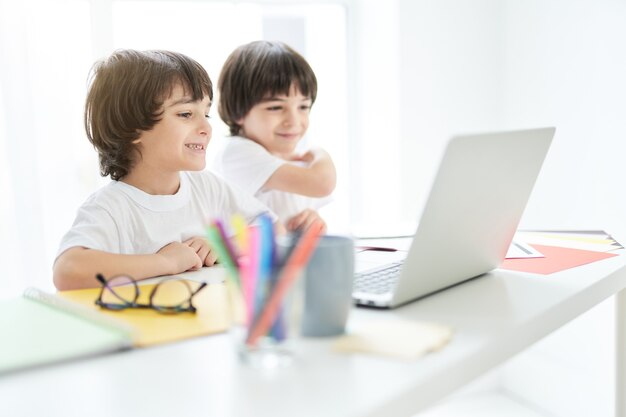 Leuke latijnse jongen die er vrolijk uitziet, zittend met zijn broer aan tafel met een laptop erop. Twee kleine kinderen die thuis online les hebben. Afstandsonderwijs tijdens lockdown concept