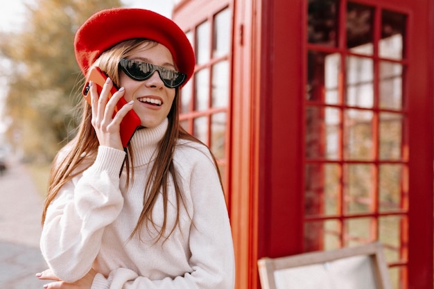 Leuke lieve vrouw met lang lichtbruin haar in witte trui en rode baret in zonnebril praat aan de telefoon en glimlacht terwijl ze opzij kijkt op de achtergrond van de telefooncel