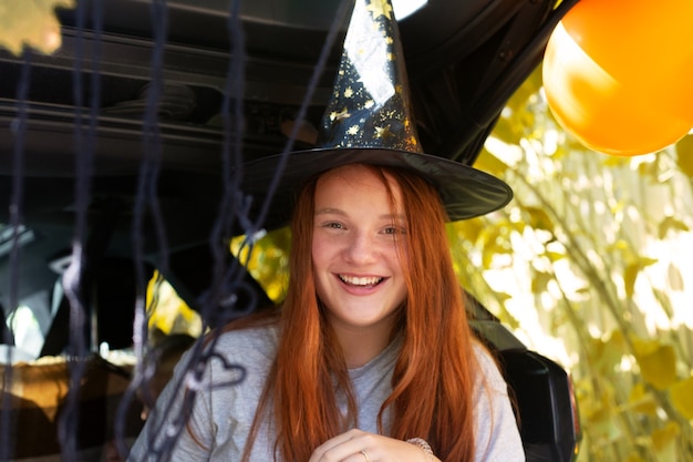 Leuke meisjesgeneratie z met lang rood haar die heksenhoed dragen die halloween in autoboomstam vieren. hoge kwaliteit foto. veilig halloween in quarantaine. snoep of je leven. herfstvakantie in isolatie