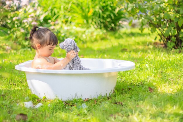 Leuke meisjeszitting in het bad met een pluchestuk speelgoed in een groene de zomertuin, op een zonnige dag
