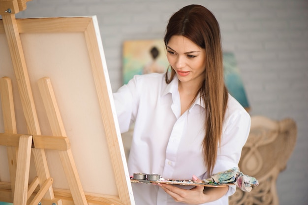 Leuke mooie meisjeskunstenaar die een beeld op een canvas op een schildersezel schildert. Studio witte achtergrond. Lang haar, brunette. Kleurrijke borstel en palet houden.