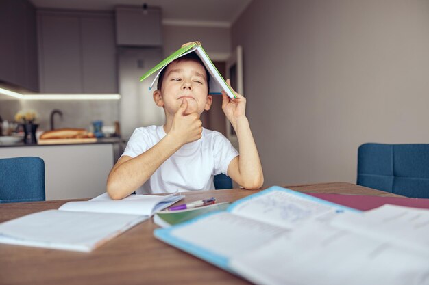 Leuke nieuwsgierige kaukasische kleine schooljongen die een leerboek boven zijn hoofd vasthoudt met een gissend gezicht