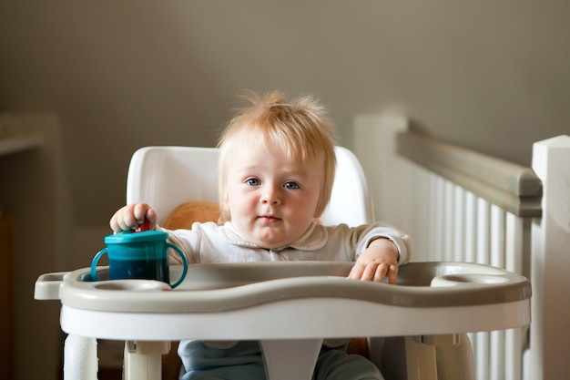 Leuke peuterjongen die in de kinderstoel zit en naar de camera kijkt met het eerste kopje van acht maanden oud