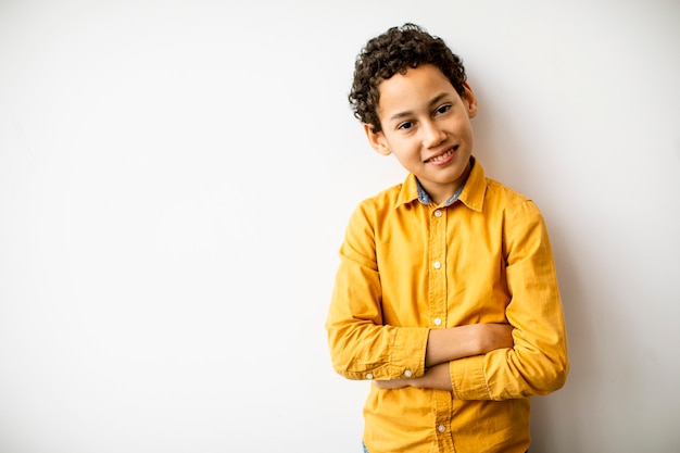 Leuke positieve kleine krullende haarjongen die zich bij de witte muur bevindt