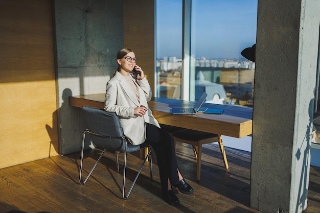 Leuke positieve vrouwelijke werknemer met lang blond haar in vrijetijdskleding die naar de telefoon kijkt terwijl ze aan een nieuw zakelijk project werkt aan tafel met laptop en gadgets op kantoor