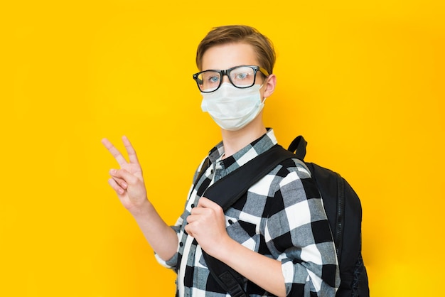 Leuke schooljongen met rugzak en gezichtsmasker geïsoleerd op gele achtergrond V, overwinningsgebaar maken. Terug naar school in het concept van de coronavirustijd