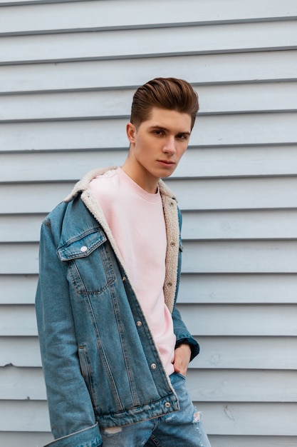 Leuke sexy jongeman met kapsel met stijlvol blauw spijkerjasje in roze modieus sweatshirt in de buurt van witte muur in de stad. Mode stedelijke lieve man in mooie trendy vrijetijdskleding wandelingen. Street Style