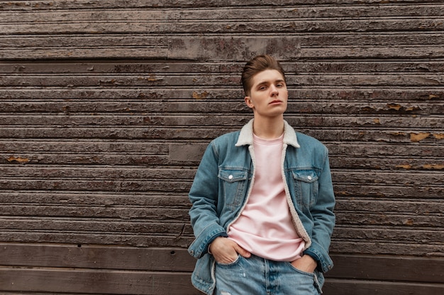 Leuke sexy jongeman met kapsel met stijlvol blauw spijkerjasje in roze modieus t-shirt in de buurt van vintage houten muur in de stad. Mode stedelijke man in mooie trendy vrijetijdskleding wandelingen. Street Style.