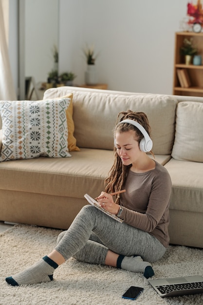 Leuke student met dreadlocks die nieuwe ideeën opschrijven in Kladblok terwijl ze huiswerk voorbereiden door de bank op de vloer