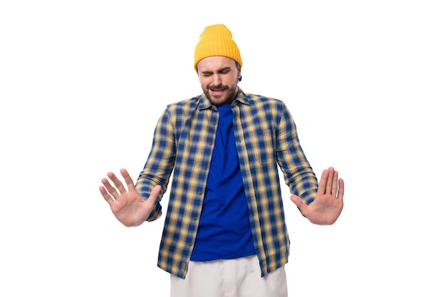 Foto leuke verbaasde jonge hipster man in gele pet en shirt werpt zijn handen op witte achtergrond