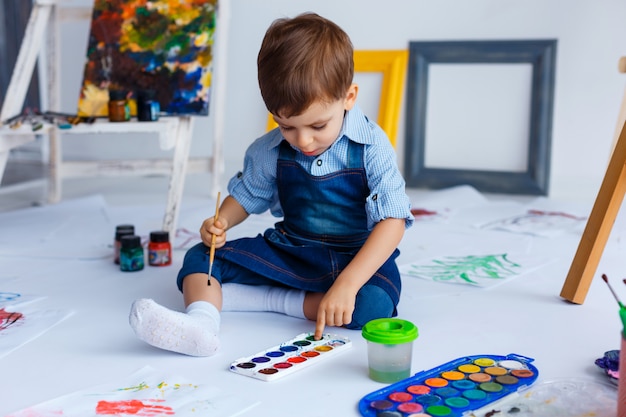 Leuke, vrolijke, blanke jongen in blauw shirt en spijkerbroek tekent met verf