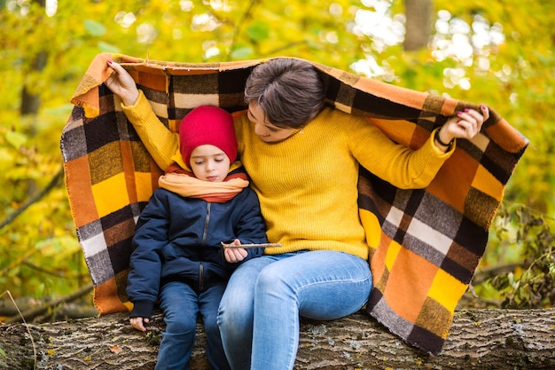 Leuke vrolijke jonge moeder is bedekt met een plaid met haar kleine charmante baby tijdens het wandelen in het herfstpark