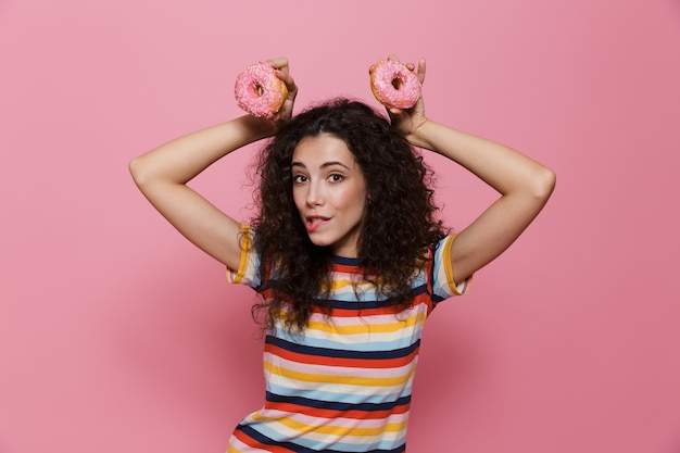 leuke vrouw 20s met krullend haar die plezier heeft en donuts vasthoudt die op roze worden geïsoleerd