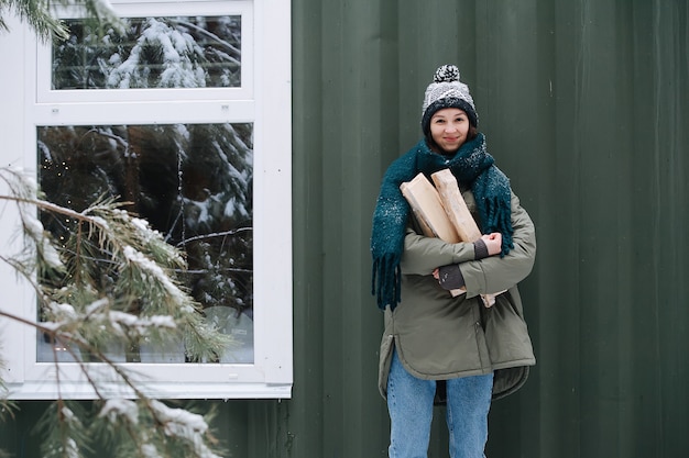 Leuke vrouw in besneeuwde winterkleren met logboeken in handen. Ze draagt een gebreide muts en sjaal. Enthousiast lachend met gesloten mond. Staande voor een huismuur.