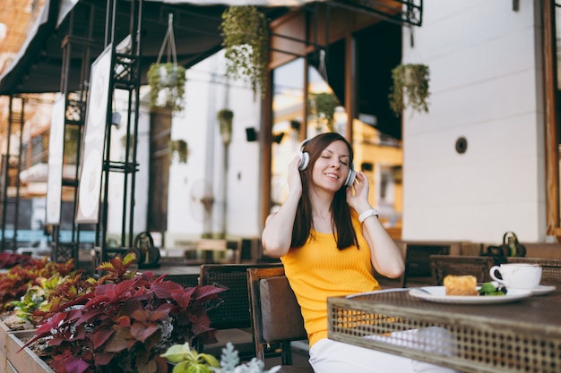 Leuke vrouw in de buitenlucht, straatcafé, zittend aan tafel in gele kleding, luisterend naar muziek in een koptelefoon