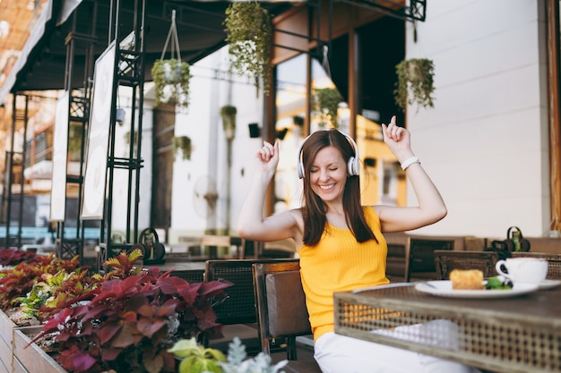 Leuke vrouw in de buitenlucht, straatcafé, zittend aan tafel in gele kleding, luisterend naar muziek in een koptelefoon