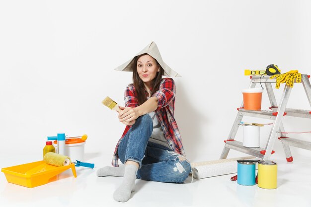 Leuke vrouw in krant hoed zittend op de vloer met borstel, instrumenten voor renovatie appartement kamer geïsoleerd op een witte achtergrond. Behang, accessoires om te lijmen, tekengereedschappen. Reparatie huis concept.