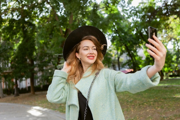 Leuke vrouw in vilten hoed en jas maakt selfie op smartphone buitenshuis