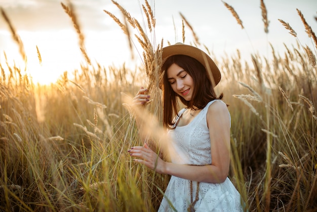 Leuke vrouw in witte jurk en strooien hoed houdt tarwe boeket in het veld. mooi meisje op de zomerweide