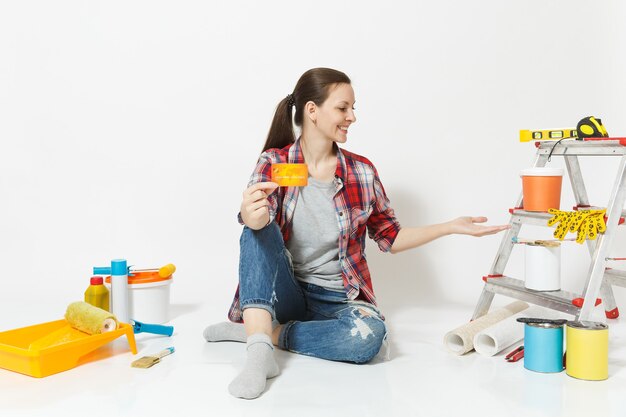 Leuke vrouw met creditcard met geld zittend op de vloer met instrumenten voor renovatie appartement geïsoleerd op een witte achtergrond. Behangaccessoires voor het lijmen van tekengereedschappen. Reparatie huis concept.