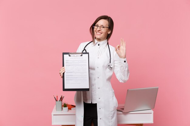 Leuke vrouwelijke arts staan voor bureau met pc-computer, medisch document in ziekenhuis geïsoleerd op pastel roze muur achtergrond. Vrouw in medische toga glazen stethoscoop. Gezondheidszorg geneeskunde concept