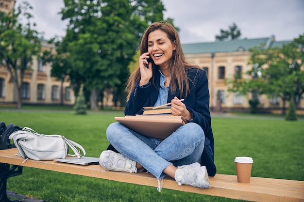 Leuke vrouwelijke student zittend op een bankje met boeken en koffie terwijl ze aan de telefoon praat en lacht