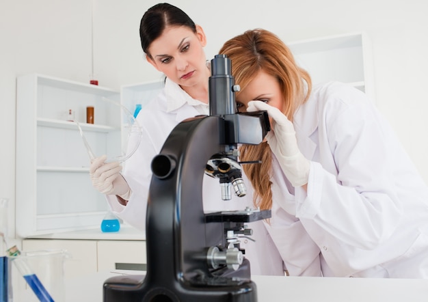 Leuke vrouwelijke wetenschapper die door een microscoop kijkt die door haar medewerker wordt geholpen