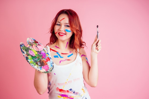 Leuke vuile make-up artist schilder vlekken van verf op het gezichtspalet in handen op een roze achtergrond in studio