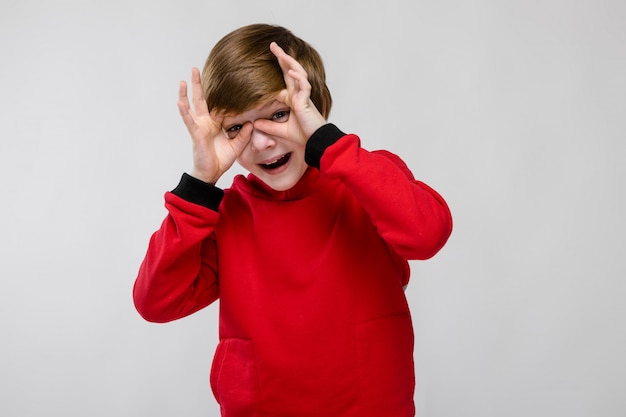 Leuke zekere Kaukasische kleine jongen in rode sweater die grappig gebaar op grijze muur tonen