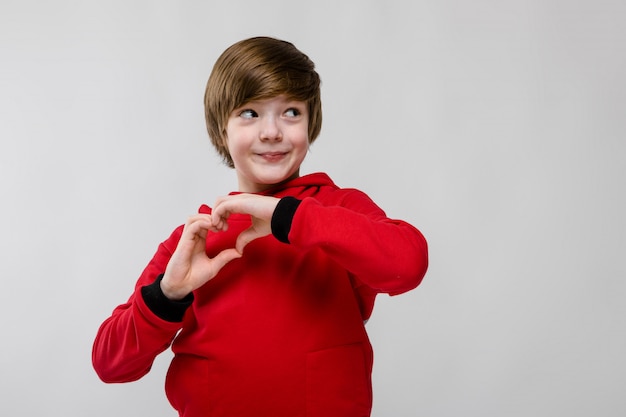 Leuke zekere opgewekte kleine Kaukasische jongen in rode sweater die liefdeteken tonen