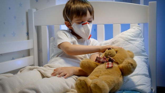 Foto leuke zieke jongen met beschermend medisch masker liggend in bed met teddybeer concept kindervirus