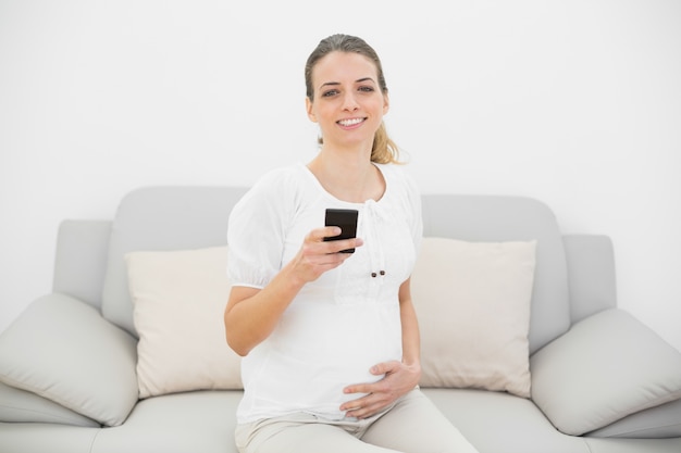 Leuke zwangere vrouw die haar smartphone houdt die gelukkig bij camera glimlacht