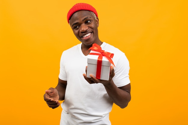 Leuke zwarte afrikaanse man met een glimlach in een wit t-shirt houdt een doos een geschenk met een rood lint voor valentijnsdag op een gele achtergrond