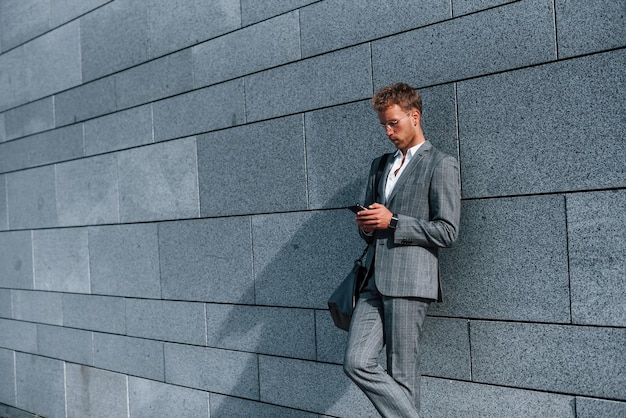 Leunt op de muur Jonge succesvolle zakenman in grijze formele kleding is buiten in de stad