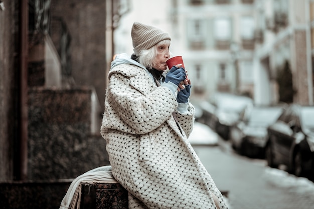 Leven in armoede. Arme senior vrouw die thee drinkt terwijl ze op straat zit