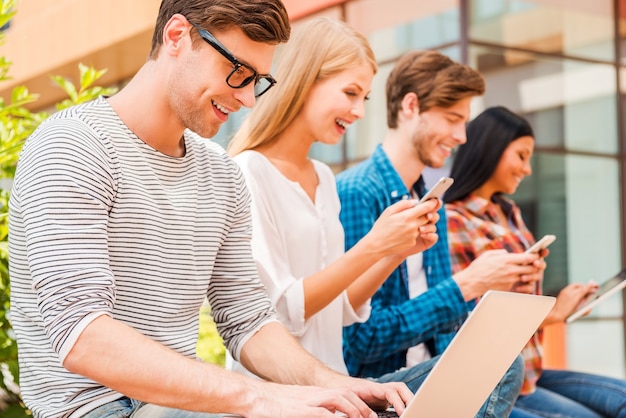 Leven in het digitale tijdperk. Groep jongeren die verschillende digitale apparaten vasthouden en glimlachen terwijl ze buiten op een rij zitten