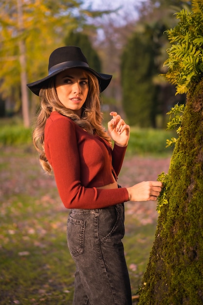 Levensstijl blonde kaukasische meisje in een rode trui en een zwarte hoed genieten van de natuur in een park met bomen portret van de jonge vrouw kijken naar de prachtige boom met mos in de herfst