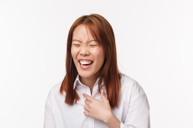 Levensstijl en mensen concept. Close-up portret van zorgeloos grappig Aziatisch meisje hardop lachen met stralende glimlach en gesloten ogen, aanraking borst grinnikt van hilarische grappen op witte muur