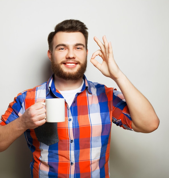 Levensstijl, geluk en mensen concept: jonge, bebaarde man met een kopje koffie in de hand en het tonen van okey, tegen een grijze achtergrond.