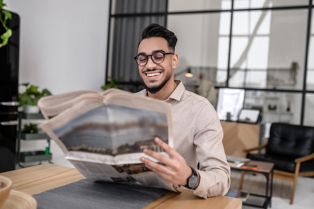 Levensstijl. Gelukkig aantrekkelijke succesvolle man in glazen glimlachend krant lezen thuis zittend aan tafel