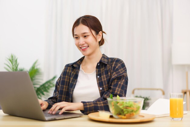 Levensstijl in woonkamerconcept Jonge Aziatische vrouw die groentesalade eet en aan laptop werkt