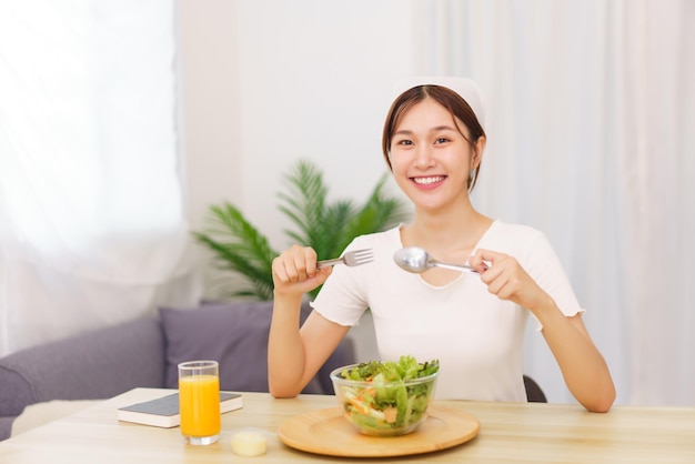 Levensstijl in woonkamerconcept Jonge Aziatische vrouw die groentesalade en jus d'orange eet